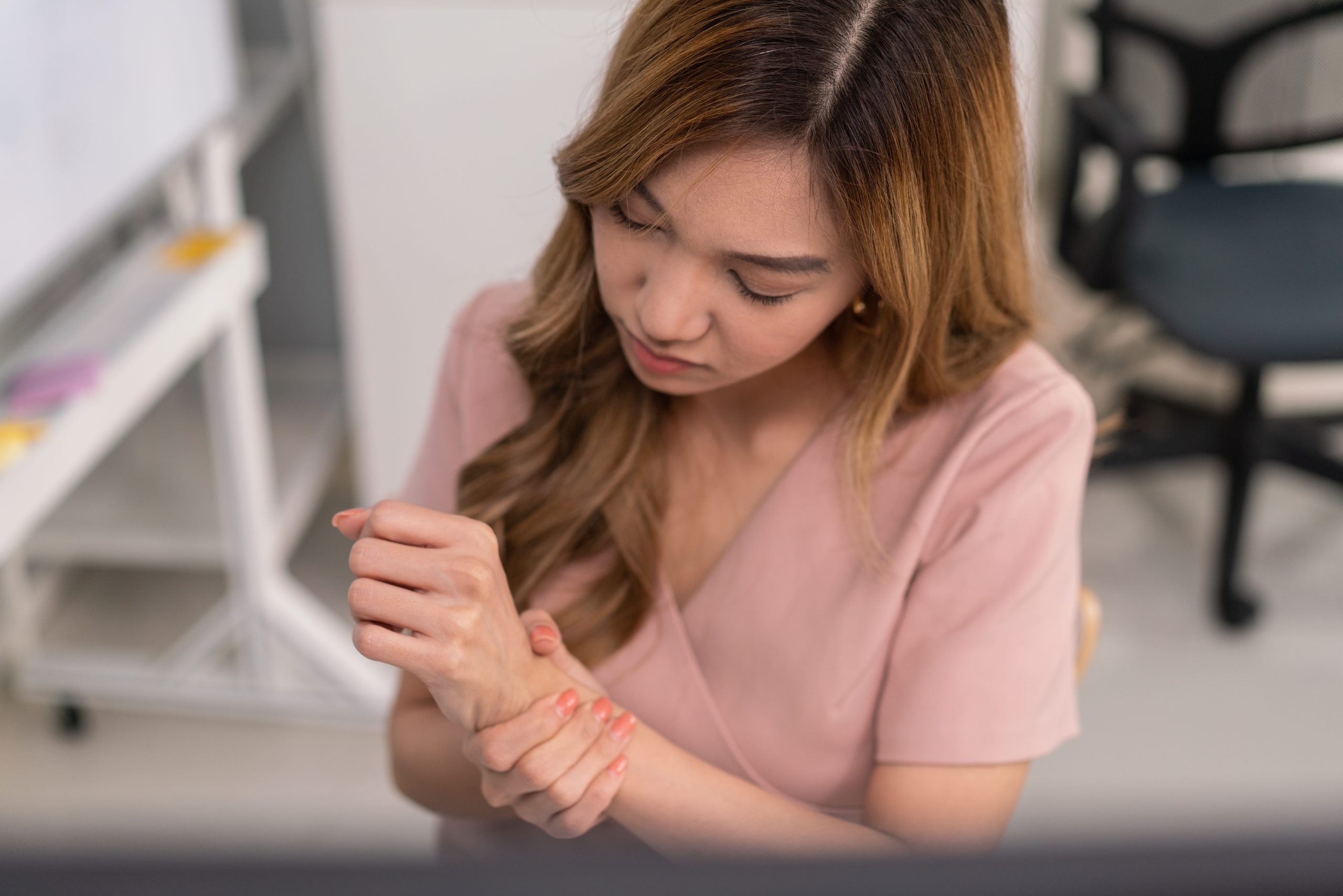 An Asian woman in a pink blouse holding her wrist in pain while looking down, indicating discomfort.